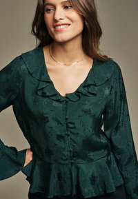 Blusa verde scuro con colletto arricciato, motivo floreale, maniche lunghe con polsini svasati e chiusura frontale con bottoni, vita peplum.