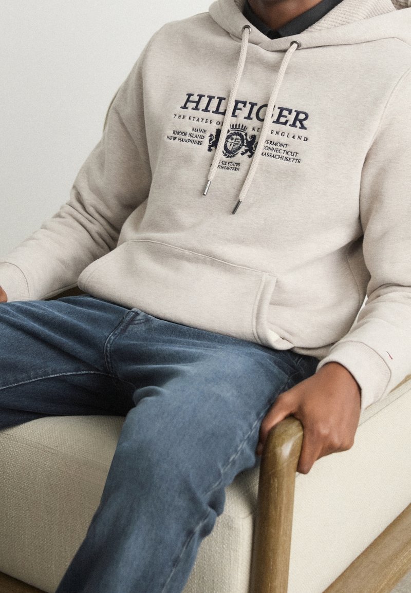 Sudadera crema con logo impreso en azul marino, capucha con cordón y bolsillo tipo canguro, combinada con jeans de mezclilla azules. Sentado en una silla de color claro.