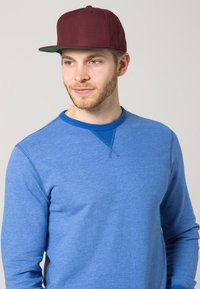 Gorra snapback burdeos con visera plana, combinada con una camiseta de manga larga azul con cuello redondo y tejido texturizado.