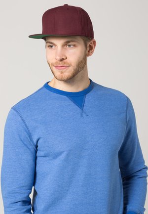 Bordo snapback kapa s ravnim vizirjem, nosi se s plavom dugom rukavom majicom s ovratnikom i teksturiranim materijalom.