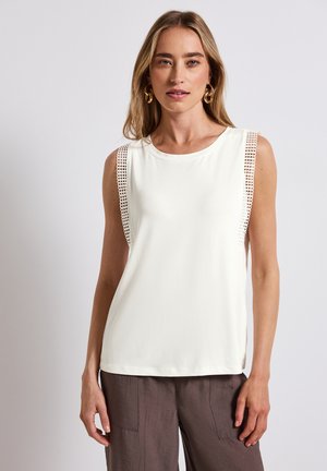 Top bianco senza maniche con una texture liscia, caratterizzato da dettagli in pizzo sulle spalle. Abbinato a pantaloni marroni larghi.