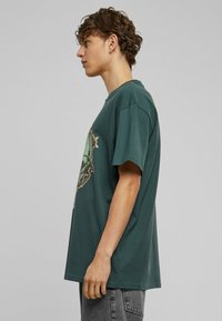 Teal bomulds T-shirt med rund hals, korte ærmer og et grafisk print på forsiden med detaljerede designelementer.
