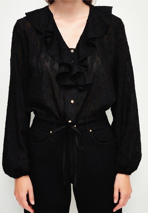 Czarna prześwitująca bluzka z falbankowym kołnierzem i złotymi guzikami, długie puszyste rękawy, wiązana w talii, noszona z czarnymi dżinsami z wysokim stanem.