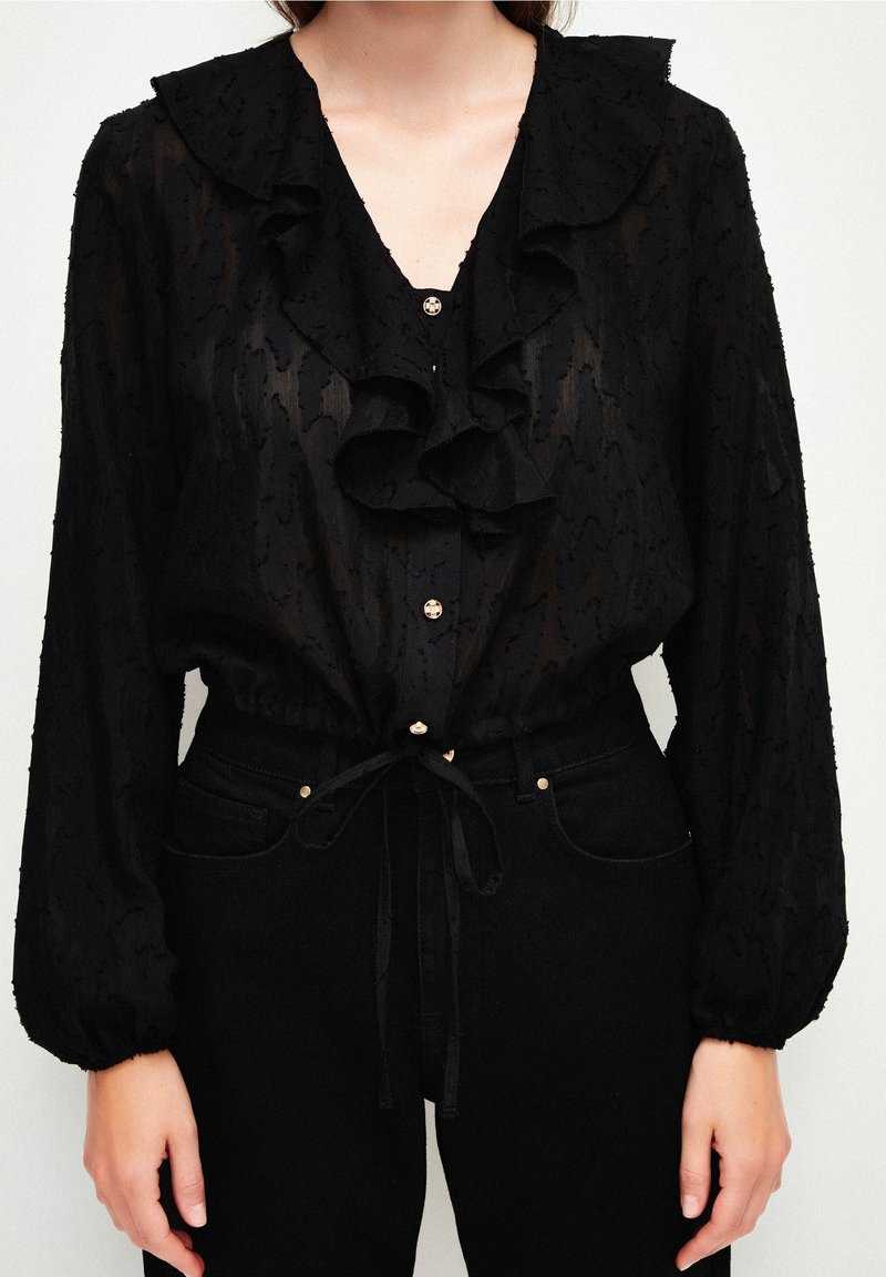 Blouse noire transparente avec col à volants et boutons dorés, manches longues bouffantes, nouée à la taille, portée avec un jean noir taille haute.