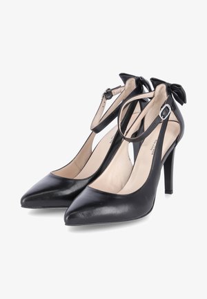 Schwarze Leder-Pumps mit hohen Absätzen und spitzen Zehen, Knöchelriemen und kleinen Schleifenakzenten an der Ferse. Glatte Textur, elegantes Design.