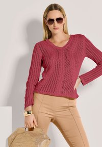Frau trägt einen rosenpinkfarbenen Kabelstrickpullover, beige Hose, große Creolen, Sonnenbrille, goldenen Armreif und hält eine geflochtene Handtasche.