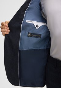 Marine blazer interieur met lichtblauwe voering, voorzien van een klein zakje en een label. Getextureerde buitenstof met een subtiel patroon.