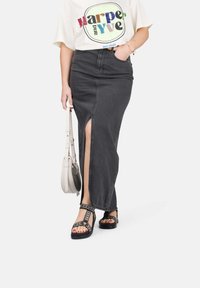 Gonna maxi in denim nero con uno spacco laterale, abbinata a una leggera t-shirt grafica e sandali neri con decorazioni. Tiene una borsa a tracolla color crema.