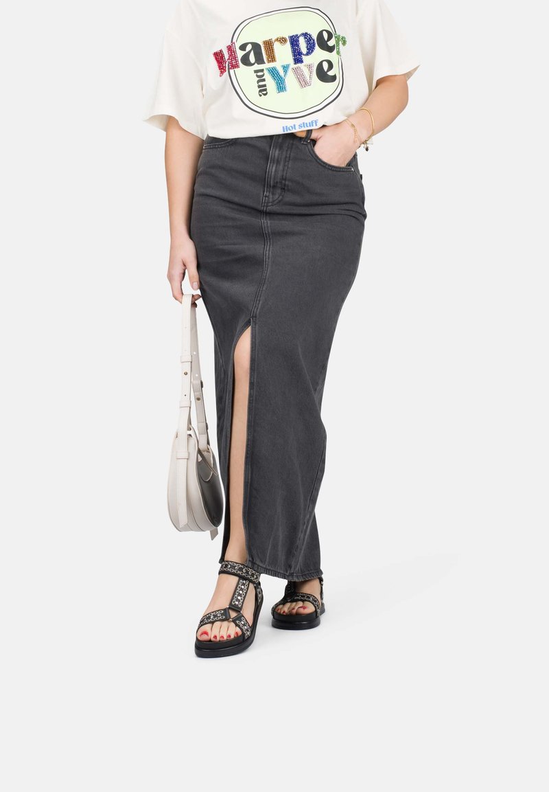 Gonna maxi in denim nero con uno spacco laterale, abbinata a una leggera t-shirt grafica e sandali neri con decorazioni. Tiene una borsa a tracolla color crema.