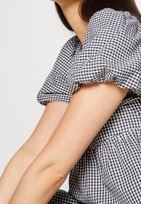 Jurk met pofmouwen in zwart-wit geruit patroon, met een geplooide halslijn en zachte textuur. Toont armen die tegen de stof rusten.