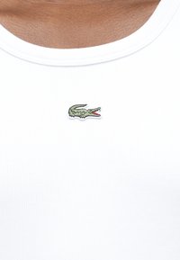 Κοντινό πλάνο ενός ατόμου που φοράει λευκό ριμπ αμάνικο με μικρό κεντημένο πράσινο λογότυπο κροκόδειλου στο κέντρο του στήθους.