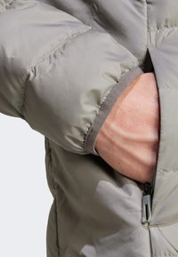 Wattierte Jacke aus hellgrauem Stoff mit strukturierter Oberfläche, elastischen Bündchen und einer seitlichen Tasche mit Reißverschluss.