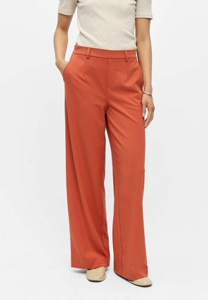 Femme portant un pantalon taille haute à jambes larges couleur rouille, top beige, chaussures beiges, une main dans la poche et l’autre reposant sur le côté.