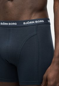 Κοντινό πλάνο ενός ατόμου που φοράει μαύρα ανδρικά εσώρουχα Björn Borg με ζώνη μέσης με το εμπορικό σήμα, εμφανίζοντας τον κορμό και μέρος του δεξιού βραχίονα.