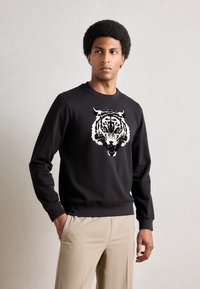 Schwarzer Pullover mit einem großen weißen Tiger-Design auf der Vorderseite. Hergestellt aus weichem Stoff mit Rundhalsausschnitt und gerippten Bündchen, kombiniert mit beigen Hosen.