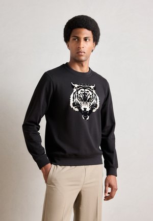 Black sweatshirt featuring a large white tiger graphic on the front. Made of soft fabric with a crew neck and ribbed cuffs, paired with beige trousers.