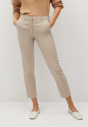 Vrouw die beige nauwsluitende enkelbroeken draagt, een geribbelde camelkleurige trui met opgerolde mouwen en witte instapschoenen met platte zolen.