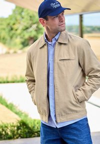 Man wearing beige jacket over blue striped shirt, blue cap with text, and jeans, standing outdoors with hands in pockets.