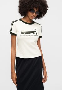 Λευκό κοντομάνικο μπλουζάκι με πράσινες λεπτομέρειες, με το κείμενο "ESPN" και ένα αθλητικό λογότυπο. Συνδυάζεται με μαύρη φούστα.