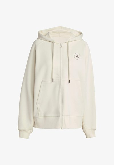 Sudadera con cremallera de color crema, hecha de un material suave. Cuenta con un bolsillo frontal, capucha con cordones y un logo circular en el área del pecho izquierdo.