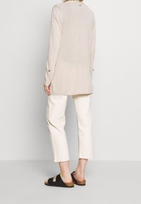 Světlý béžový dlouhý pletený kardigan s žebrovaným lemem a knoflíkovými manžetami, doplněný o krémové krátké kalhoty a černé pantofle na klínku.