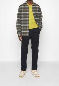 Rutig grå och vit skjorta över en klar gul t-shirt, kombinerad med mörka byxor och beige sneakers med vita detaljer.