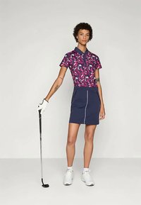 Kvinna i mönstrad polotröja, marinblå kjol, vit golfhandske och vita sneakers håller en golfklubba i en neutral studio miljö.