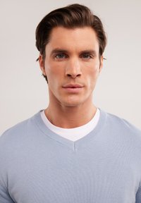 Suéter de punto fino en azul claro con cuello en V, superpuesto a una camiseta blanca de cuello redondo. Textura suave con un sutil detalle en el tejido.