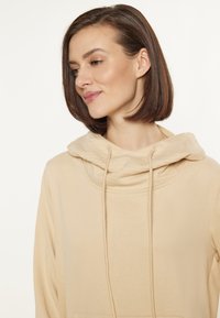 Beige kapucnis pulcsi, húzott nyakkal és zsinórokkal. Simma textúrával és elülső zsebből készült. Egyszerű és minimalista kialakítás.