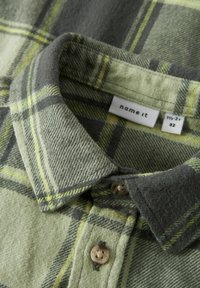 Grønn rutete flanellskjorte med knapp ned-krage, beige knapper og et teksturert stoff med gule og grå aksenter. Merkeetikett synlig.