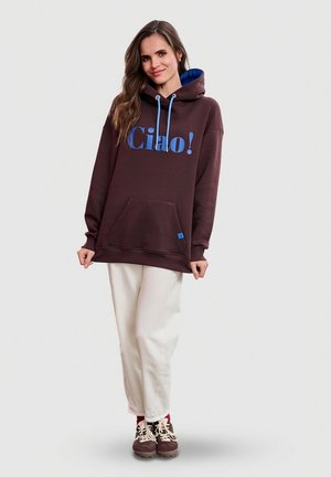 Brązowa, oversize'owa bluza z kapturem z napisem "Ciao!" w kolorze niebieskim, z niebieskim podszyciem kaptura, przednią kieszenią i ściągaczami na rękawach. Noszona z białymi spodniami i brązowymi butami.