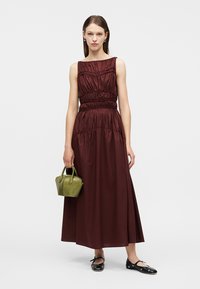 Γυναίκα με μακρύ μαν sleeveless μαρούλι σε μπορντό, κρατώντας μια μικρή ελιά πράσινη τσάντα χειρός, φοράει μαύρα μπαλαρίνια με ασημένια καρφιά.