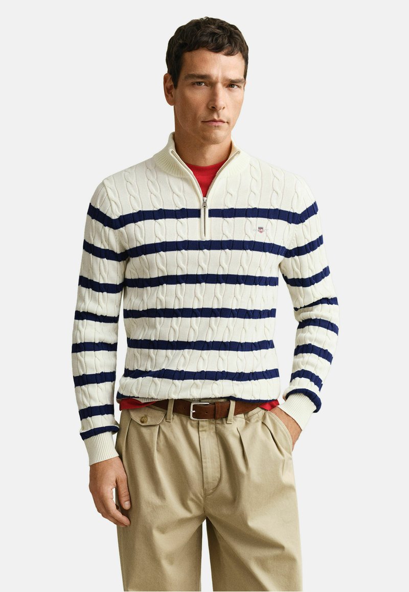 Homme portant un pull blanc en maille torsadée avec des rayures horizontales bleu marine, une chemise rouge en dessous, un pantalon plissé beige et une ceinture marron, debout la main dans la poche.