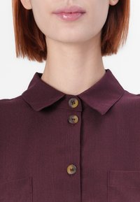Burgundy skjorta med klassisk krage, två knappar vid halsen och en ficka på vänster sida. Slät textur, skräddarsydd passform.