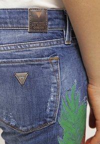 Modré džíny z denimu s opotřebovaným vzhledem. Obsahují hnědou koženou našivku, trojúhelníkové logo a výšivku zeleného listu na kapse.