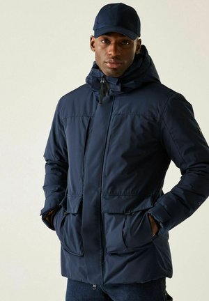 Mężczyzna ubrany w granatową zimową kurtkę z kapturem i przednimi kieszeniami z klapami oraz granatową czapkę z daszkiem, ręce w kieszeniach, neutralne tło.