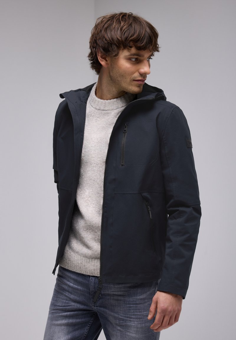 Veste imperméable bleu marine foncé avec capuche, fermeture éclair à l'avant et plusieurs poches zippées. Portée sur un pull gris clair, associée à un jean.