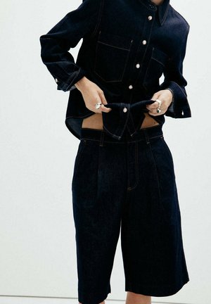 Personne portant une chemise en denim foncé et un short long taille haute assorti, les mains ajustant l'ourlet de la chemise, fond clair uni.