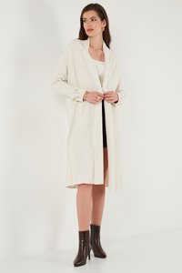 Lange, crèmekleurige ribgebreide cardigan met open voorkant, twee zakken en knoopdetails op de mouwen, gedragen met een getailleerde zwarte jurk en bruine enkellaarsjes.