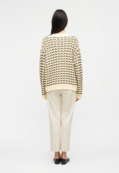 Γυναίκα που φαίνεται από πίσω, φοράει πουλόβερ πλεκτό με εκρού και μαύρο σχέδιο, εκρού παντελόνι κανονικής γραμμής και μαύρα παπούτσια με επίπεδη σόλα.