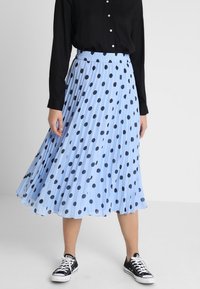 Gonna plissettata azzurro chiaro con pois neri, fascia elastica in vita, indossata con una maglietta nera e sneakers nere. Tessuto dalla consistenza liscia.