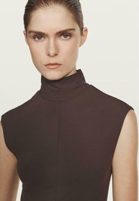 Haut marron sans manches avec un col montant, confectionné dans un tissu lisse, présentant une couture au centre et une coupe ajustée.