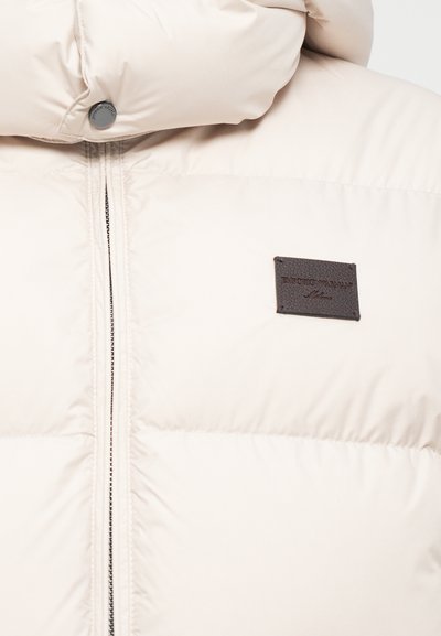 Beige pufferjakke med høj krave, der har en frontlynlås og et logo-patch i brunt læder. Glat, kviltet tekstur.