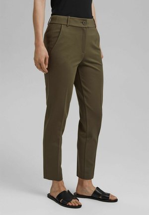 Kvinde iført olivengrønne skræddersyede ankellange bukser og sorte slidse-sandaler, stående mod en ensfarvet grå baggrund.