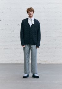 Oversized dark cable-knit cardigan, white collared shirt with a bow tie, light gray wide-leg trousers, and black athletic shoes with teal accents.