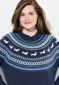 Donna con lunghi capelli castani che indossa orecchini a cerchio dorati e un maglione blu navy decorato con silhouette di cani bianchi e disegni geometrici.