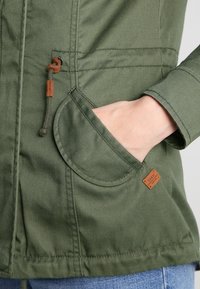 Olivgrön jacka med texturerad tyg, rund ficka, justerbar dragsko i midjan och ett märkt läderpatch.