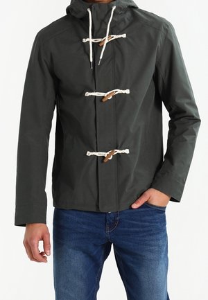 Musta huppari, jossa kolme valkoista köysi- ja puunappisulkemista, puettu sinisten farkkujen kanssa, vasen käsi taskussa.