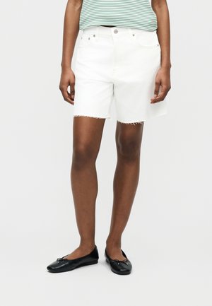 Inimene, kes kannab valgeid teksapükse franjaliste äärtega, triibulist rohelist ja valget t-särki ning musti baleriinijalatseid, seisab ühevärvilise tausta ees.