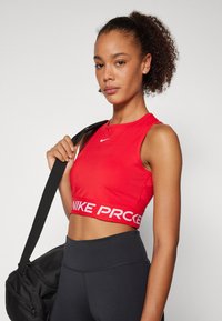 Crvena skraćena majica bez rukava s bijelim „NIKE PRO“ remenom. Glatka tekstura i dizajn bez rukava. Model drži crnu torbu za teret. Crne uske tajice.